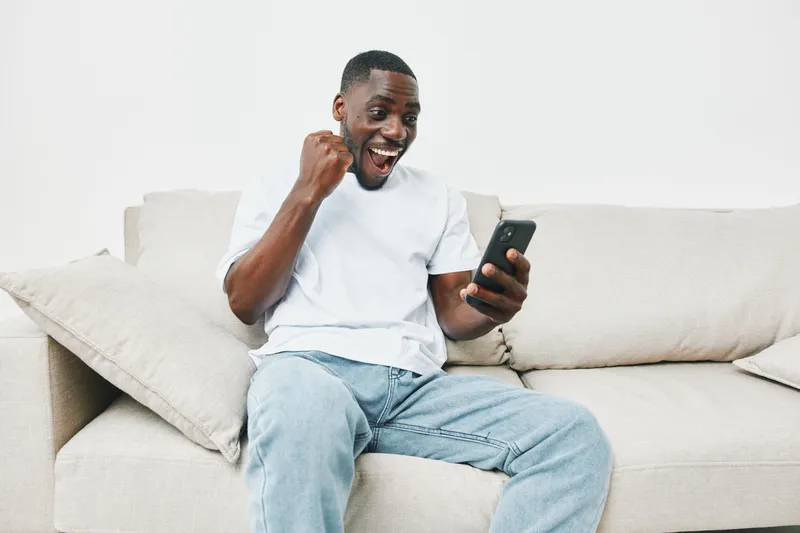 Excited African American man celebrating success while checking his phone on a cozy couch, showcasing emotion and joy in a modern living room setting