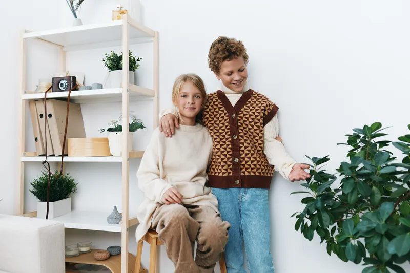 Two young siblings share a warm moment indoors, one sitting on a chair, the other smiling beside a lush houseplant, in a bright, minimalist living space.