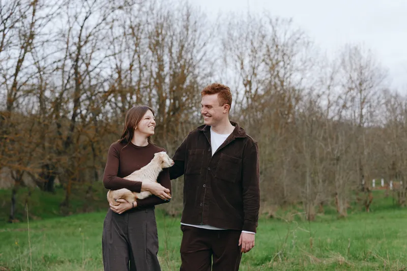 A joyful couple walks outdoors with a goat kid in a grassy park, sharing smiles and gentle interaction. A wholesome lifestyle moment in nature, showcasing companionship and animal bonding.