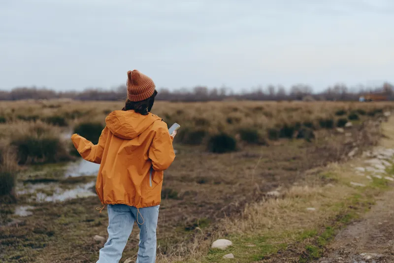 Woman outdoors lifestyle, walking in a rural field, wearing a yellow hooded jacket, seen from behind, calm nature background, cloudy sky, relaxed weekend mood, soft breeze, open air, today