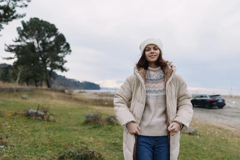 Woman in a beige puffer coat and knit sweater outdoors by a quiet coast, grassy field, cloudy sky, and distant parked car behind her for a calm winter moment