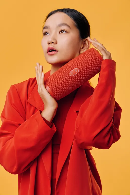 Woman in red jacket holding portable speaker close to face against orange background for music, lifestyle, technology and sound concept with casual style and modern gadget.