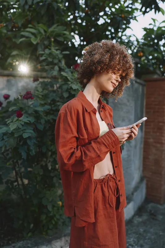 Cheerful woman outdoors, focused on her smartphone, casual rust outfit, warm sunlight, leafy background, relaxed posture, contemporary lifestyle moment, soft light