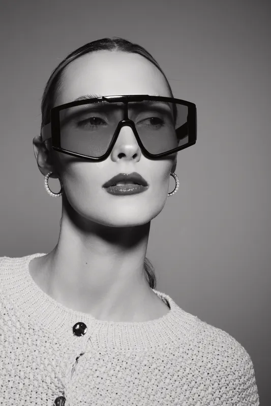 Young white woman with light skin wearing large sunglasses and knitted sweater looks aside. Studio portrait in black and white with neutral background.