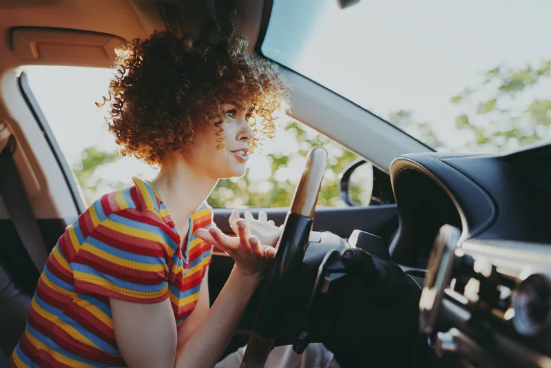 Curly haired woman driving a car wearing a striped tshirt smiles as sunlight fills the cabin, creating a warm casual mood that highlights everyday travel and relaxed confidence today.