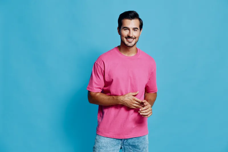 Smiling young man wearing a pink t-shirt stands against a blue background, looking at the camera with a friendly expression. Casual lifestyle concept.