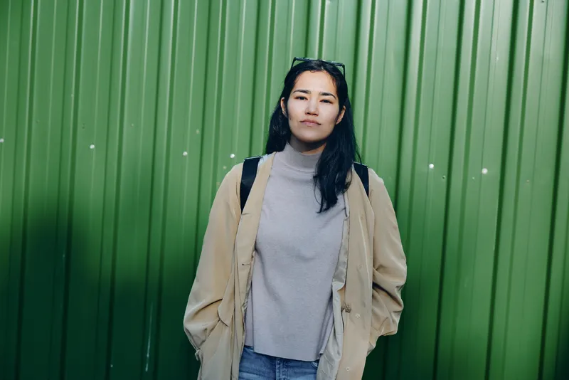 Woman wearing a beige coat over a gray turtleneck stands against a vibrant green wall, casual street fashion, confident pose, urban backdrop and calm mood.