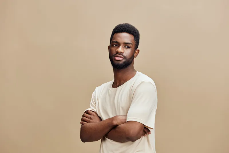Confident young black man with crossed arms in a neutral beige background showcasing self assurance and style
