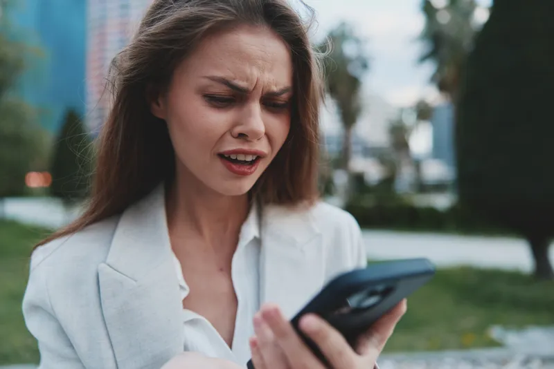 Frustrated woman in white jacket looking at smartphone stressed by financial crisis and stock market decline outdoors in spring season