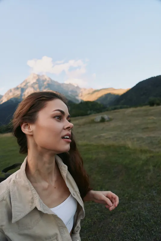 A young woman stands on a grassy hillside with distant mountains, wearing a beige shirt over a white top, looking to the side with a contemplative expression.