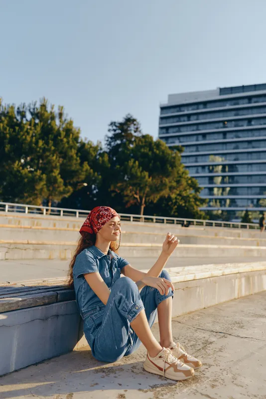 relaxed woman sitting outdoors wearing denim jumpsuit and red bandana enjoying warm sunshine with urban park background in bright daylight