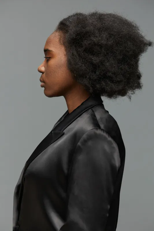 Profile portrait of a young Black woman with natural hair, wearing a black blazer on a neutral gray background, symbolizing confidence and self expression