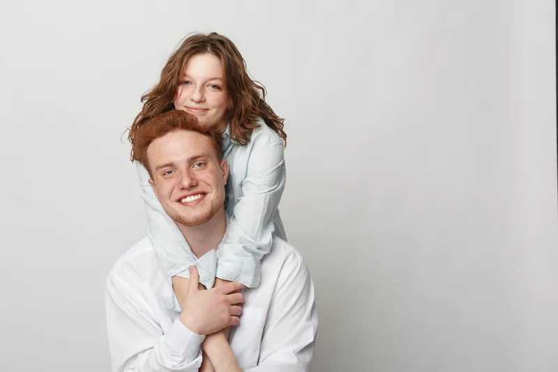 Smiling young man with red hair carries happy girl on his back. Both wear white shirts and pose against plain light background. Concept of family, friendship, and joyful moments.