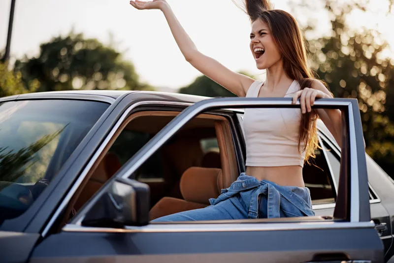 Joyful young woman enjoying a sunny day in a car, waving from the window with a vibrant smile and casual summer attire, exuding happiness and freedom.