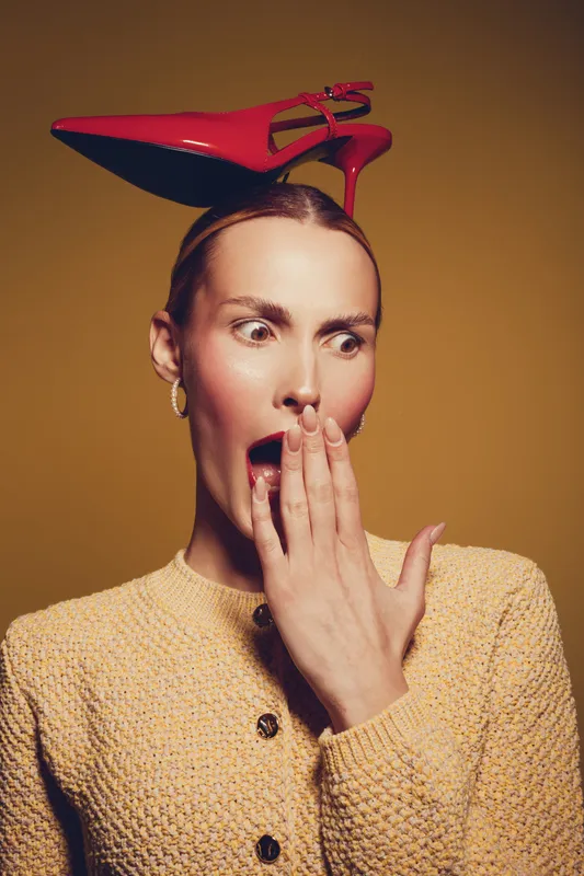 Surprised woman with red high heel shoe on head covers mouth with hand, wearing yellow textured sweater. Concept of fashion, shock, and unusual style on brown background.