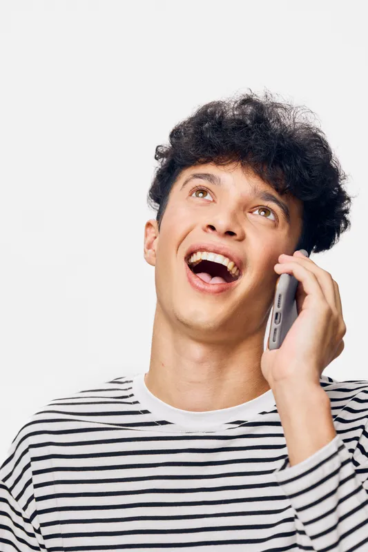 Young man talking on phone smiling joyfully with curly hair and striped shirt, isolated on white background, expressing happiness and communication.