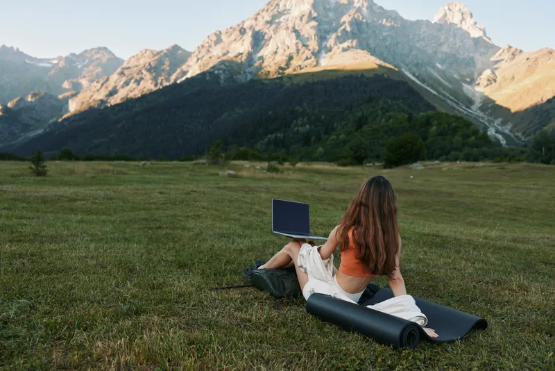 A person sits on a grassy meadow with a laptop, distant mountains in the background, creating a peaceful outdoor work moment and a sense of calm focus for creative tasks and learning.