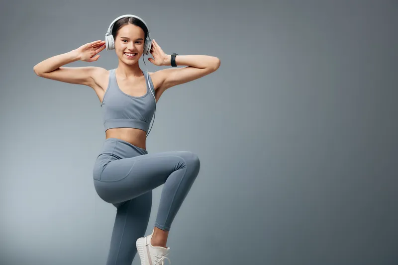 Woman fitness athlete wearing gray athletic wear and headphones, standing on one leg in a simple studio backdrop; dynamic exercise, focused and upbeat, showcasing energy and motivation in a clean,