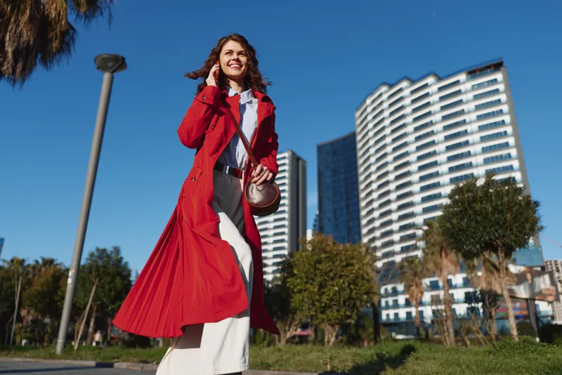 Young woman in a red trench coat walks through an urban park with tall buildings in the background, smiling and confident, showcasing bold street style.