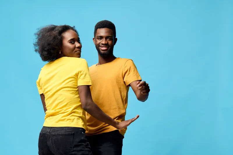 Young African couple dancing together in vibrant yellow outfits against a bright blue background, showcasing joy and connection in a playful atmosphere