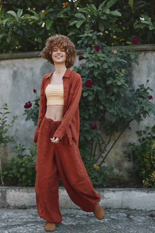 Vibrant woman lifestyle scene shows a smiling young woman wearing rust colored pants and a pale yellow crop top walking along a garden wall outdoors with casual confidence