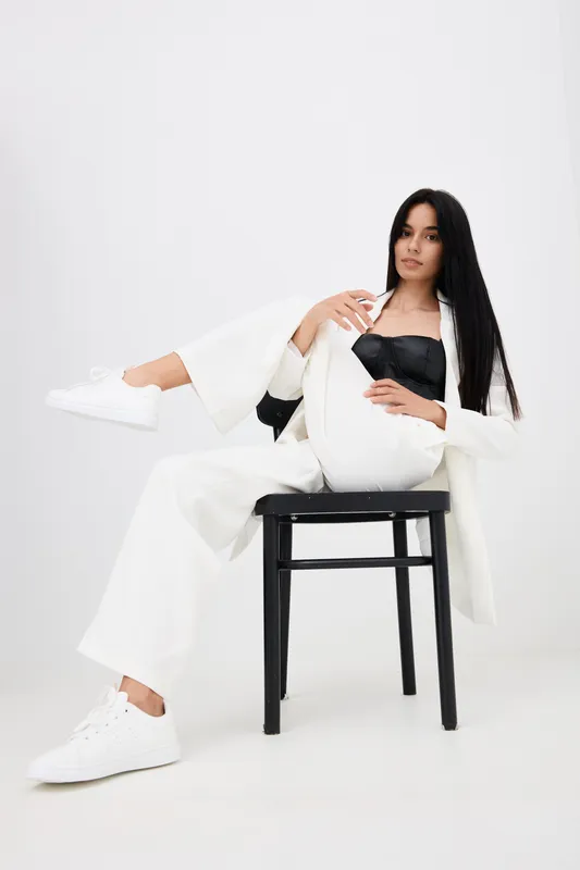 stylish woman in white suit and black top sitting with one leg on black chair, long dark hair, white sneakers, minimalist bright background