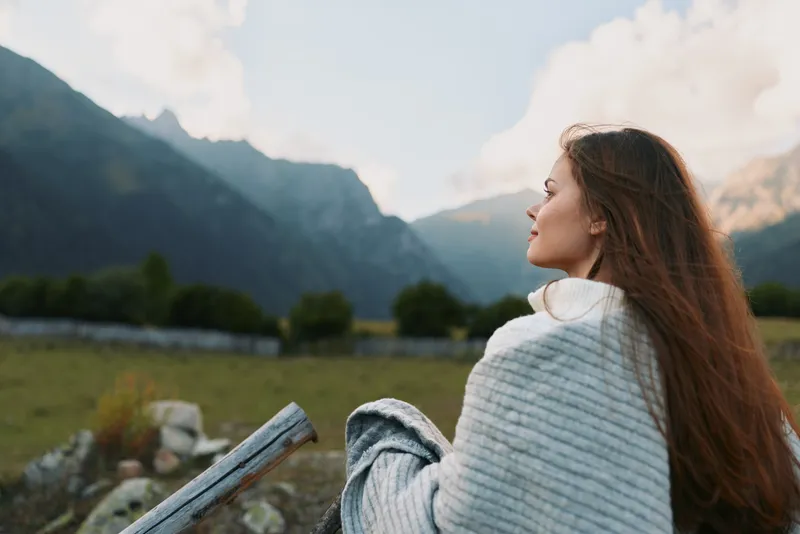 Young woman sits on the grass wrapped in a blanket, gazing toward distant mountains, conveying peaceful solitude and reflection.
