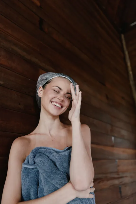 smiling woman wrapped in towel with shower cap on head stands against wooden wall indoors, expressing joy and relaxation after bath or spa session.