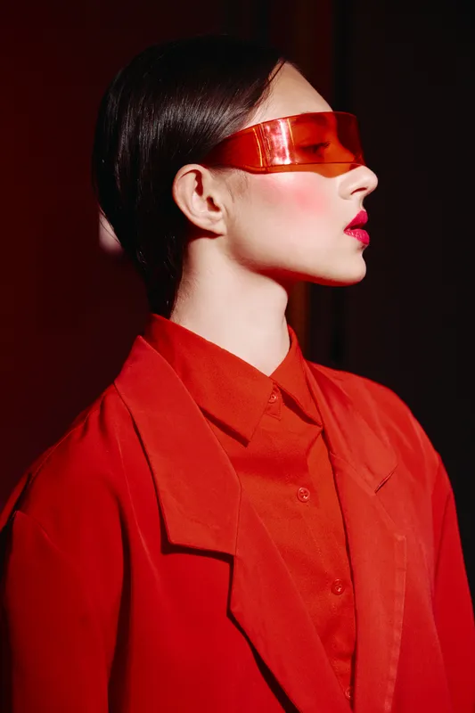 Profile of a stylish young woman with short hair wearing futuristic orange visor and red jacket against a vivid red background. Concept about fashion, beauty, and avant-garde style.
