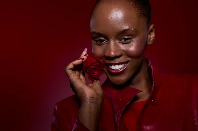 Smiling dark skinned African woman holds red rose near face in red leather jacket. Studio portrait with burgundy background. Concept of beauty, happiness, and floral elegance.