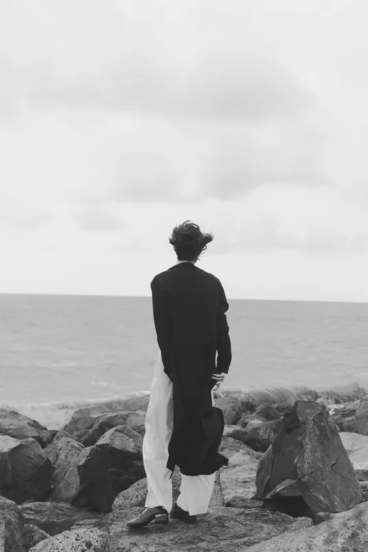 Solitude on the coast as a woman stands on rocks facing the sea, wind in her coat and flowing dress, capturing a quiet moment of contemplation and distant horizons