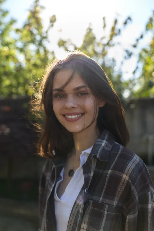 Young woman with shoulder length dark hair smiles softly in a sunlit garden, wearing a plaid shirt over a white top, creating a casual lifestyle vibe