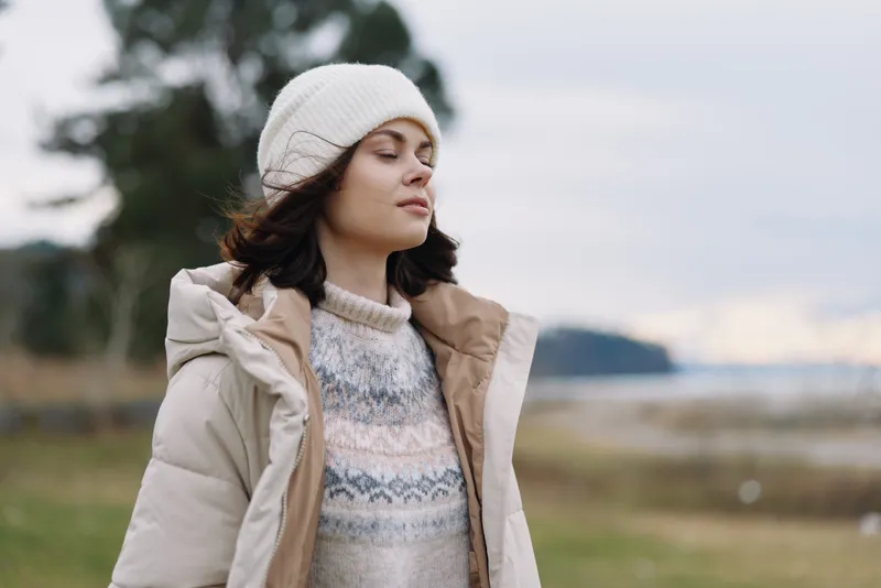 Woman outdoors in a beige winter coat, knit sweater and white beanie, eyes closed, enjoying a cold breeze in an expansive landscape with soft clouds and distant water.