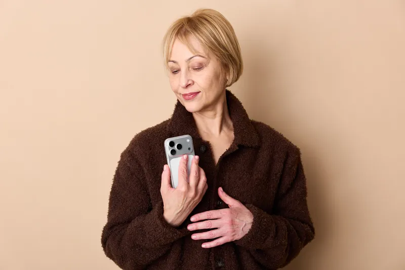 Woman with smartphone looking at screen, technology and communication concept, portrait indoors on beige background, wearing dark robe, soft studio lighting, selfcare mood