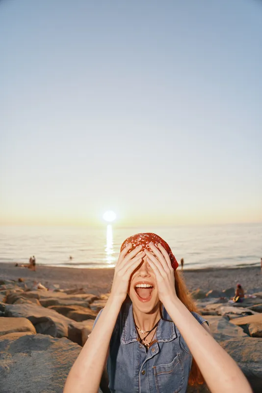 lifestyle woman covering eyes with hands in modern boho-western denim street style, enjoying sunset by the beach in film color tones