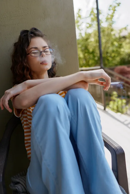 A pensive woman sits on a balcony ledge in casual home attire, blue pants and striped top, eyes closed, smoke curling from a cigarette. A quiet moment of solitude and routine.
