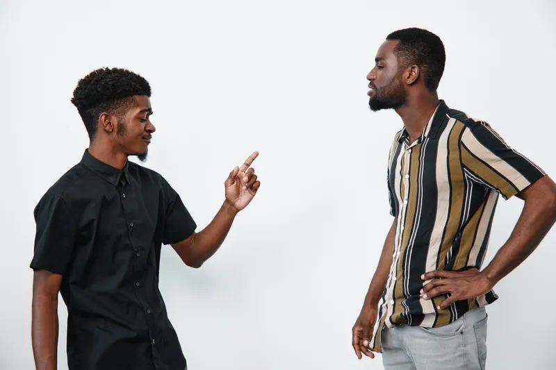 Two black men with dark skin having a serious conversation in casual clothes on white background. One man gestures while the other listens with hands on hips.