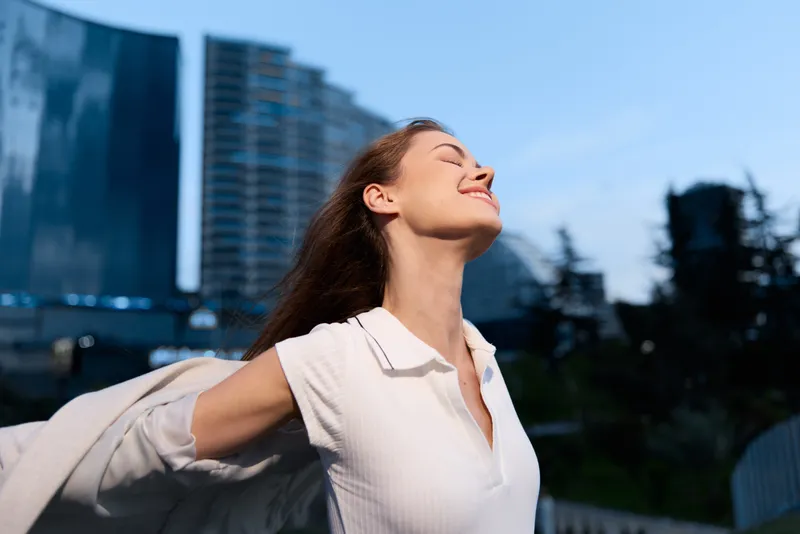 Woman feeling joyful outdoors with arms outstretched in white shirt against urban blue sky background, enjoying fresh air and freedom with happy expression