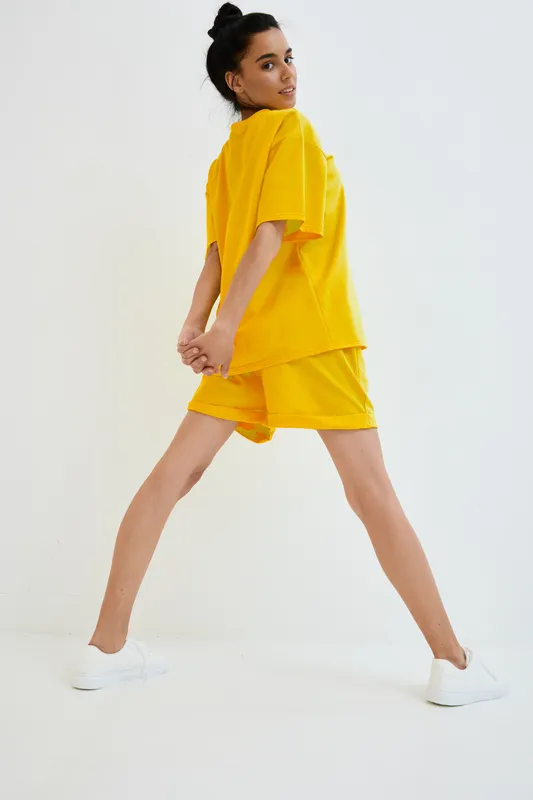 Young woman stretching in vibrant yellow sportswear and white sneakers against a plain white background, showcasing fitness and casual style in bright colors.