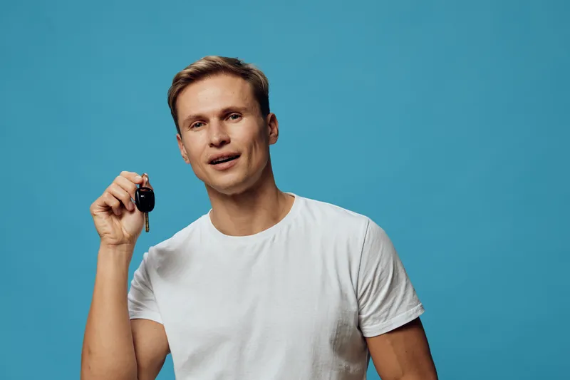 Young Caucasian man in casual white T-shirt holding car key remote isolated on blue background studio portrait. People lifestyle and transportation concept.