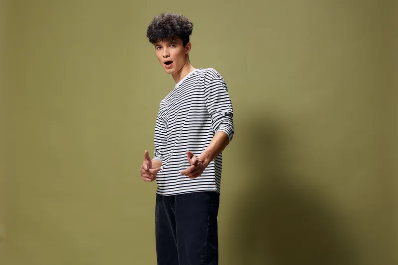 young man in striped shirt posing confidently against green background with curly hair and casual black pants, expressing surprise and engagement