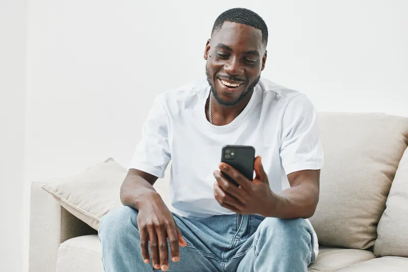 Happy Black man smiling while using smartphone indoors on a comfortable sofa, showcasing modern culture and technology in a bright and minimalistic setting