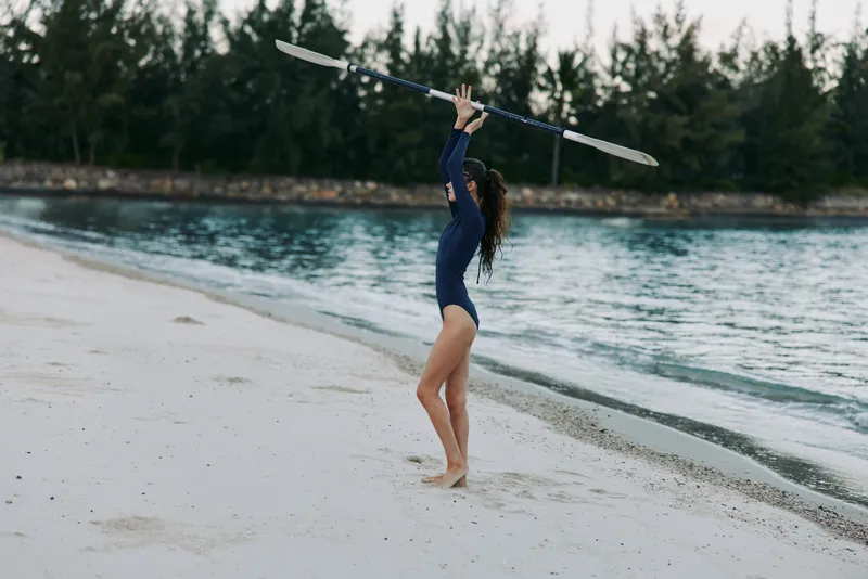 Woman in blue swimsuit holding kayak paddle on sandy beach near turquoise water under cloudy sky.