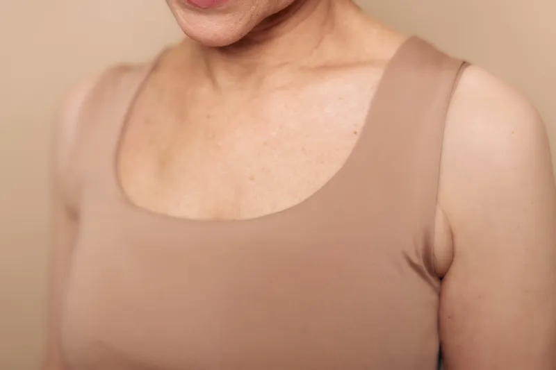 Close-up of mature woman wearing beige sleeveless top showing natural skin texture and wrinkles, soft lighting highlighting age and beauty. Concept of aging, skin care, and natural beauty.