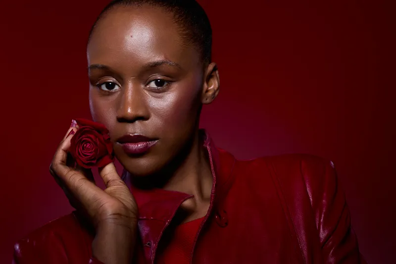 Close-up of a dark skinned African woman holding a red rose near her face wearing a red leather jacket. Concept of beauty, elegance, romance, and fashion with a red background.