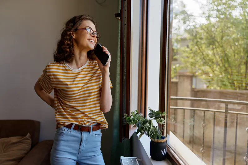 Smiling woman in a striped shirt standing by a sunlit window, enjoying a calm home moment. Casual lifestyle scene emphasizing comfort, light, and everyday joy.