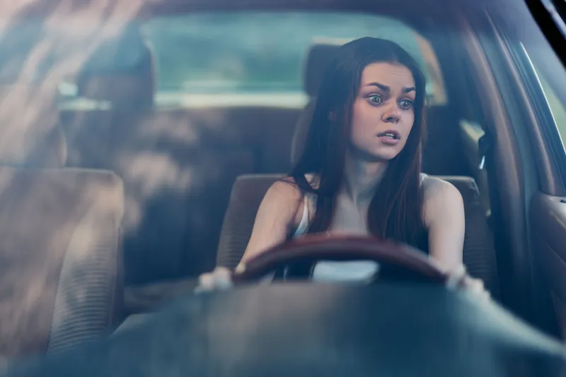 Young woman with long dark hair looking shocked while driving a car, conveying surprise and concern in a blurred natural background.