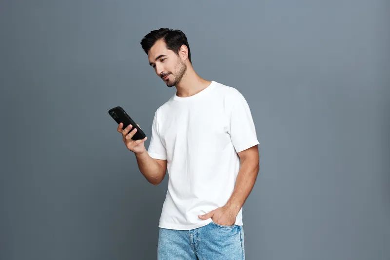 Stylish young man in casual white t-shirt standing with hand in pocket looking at smartphone, isolated on plain gray background. Modern lifestyle concept.