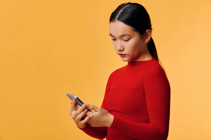 Young woman in red sweater using mobile phone for texting or browsing online on orange background. Female person focused on smartphone communication or social media app.