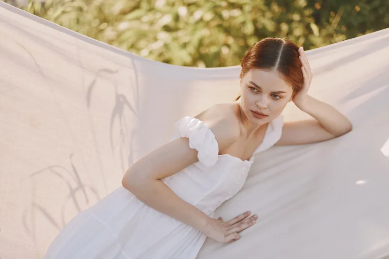 A serene young woman in a white flowy dress lounging against a light fabric backdrop, conveying tranquility in a sunlit, natural setting